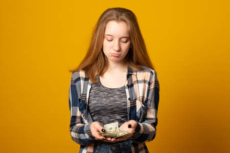 Teenage girl in casual clothes counts the first money earned. Upset young girl holding money on a yellow background.の写真素材