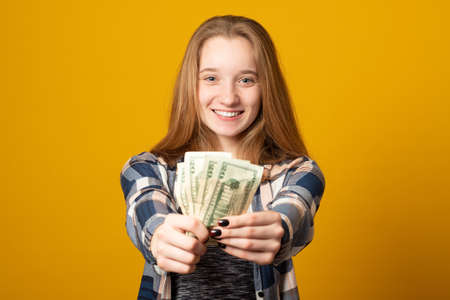 Portrait of a cheerful young girl holding money banknotes and celebrating on yellow background. Girl teenager is happy with the first dollars earned.の写真素材
