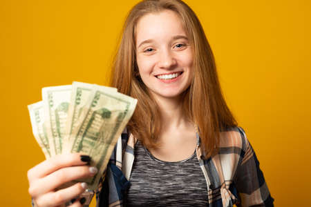 Portrait of a cheerful young girl holding money banknotes and celebrating on yellow background. Girl teenager is happy with the first dollars earned.の写真素材