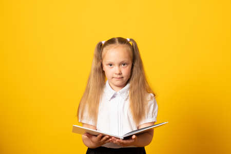 Cheerful young blonde child girl  holding a book to study isolated on yellow background