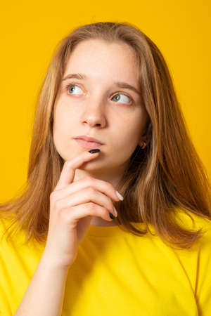 Pleasant looking teen girl holds chin and looks pensive away, thinks about great plan, has new ideas in mind, wears bright yellow t shirt.の写真素材