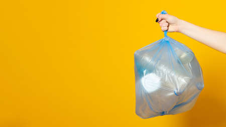 Woman hand holding blue plastic garbage bag, packet with domestic waste while on yellow background,...