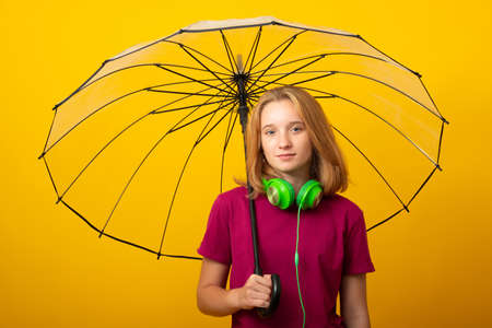 Positive happy young girl in green headphones, dressed in a lilac T-shirt, holds an umbrella on a yellow background. Bright autumn concept.の写真素材