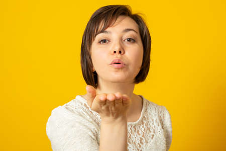 Charming european woman sends air kiss at camera, expresses love, poses against yellow studio...
