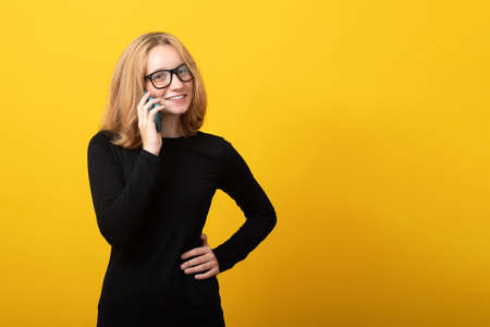 Happy young woman blonde, in glasses, dressed in a strict black dress, talking on a mobile phone, on a yellow background. Banner copy space.の写真素材
