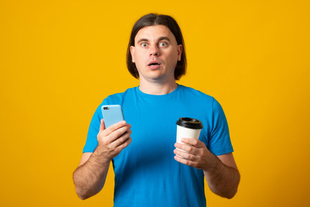 Surprised handsome man uses mobile phone for online communication and surfing internet holds takeaway coffee being in good mood dressed casuallyの写真素材