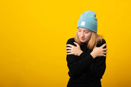 Studio portrait of young cute blonde girl embracing herself against yellow background, wearing blue hat and black sweater. Copy space.の写真素材