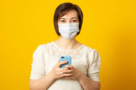 Satisfied woman has flu, wears protective medical mask not to infect other people, uses cell phone for surfing internet, reads how to cure disease online, dressed in fashionable clothes.の写真素材