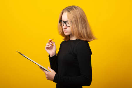 Smiling young blond business woman in black dress, glasses isolated on yellow background. Achievement career wealth business concept. Writing notes in notebook, looking aside.の写真素材