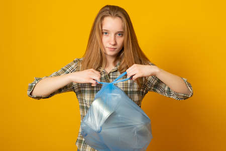 Cute young girl holding plastic trash bag on yellow background. Waste sorting and sustainability concept.の写真素材