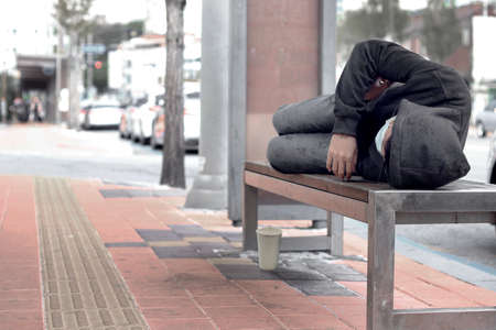 Poor homeless man or refugee sleeping on the wooden bench on the Sidewalk in the cityの写真素材