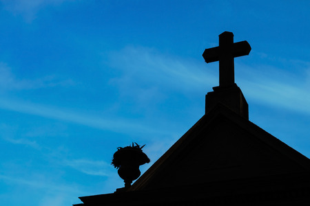 A backlit cross on the top of a cemetery mausoleum with a blue sky on the backgroundの写真素材