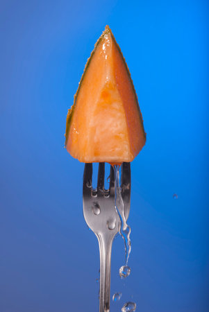 Melon fruit piece on fork with water splash and empty blue background concept photo. Ideal for healthy eating campaign, motivation or fitness poster.の写真素材