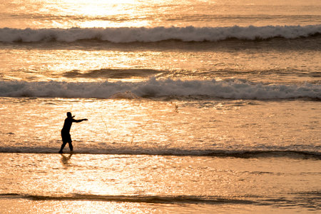 Fisherman throwing fish net on beach coast, summer calm scene at sunset time with golden sunlight on waves.の写真素材
