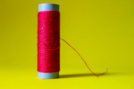 Isolated photo of a roll or a spool of red thread above and in front of a yellow background. This thread is a bit loose from the reel.の写真素材