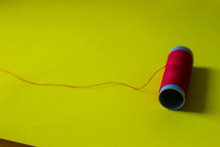 Isolated photo of a roll or a spool of red thread above and in front of a yellow background. This thread is a bit loose from the reel.の写真素材