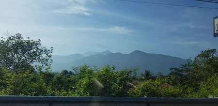 A view of green rice fields and gardens with blue sky and white clouds. You can see the silhouette of a mountain behind him. This view is the view when on the way to Yogyakarta.の写真素材