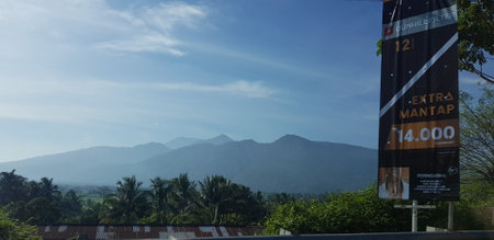 A view of green rice fields and gardens with blue sky and white clouds. You can see the silhouette of a mountain behind him. This view is the view when on the way to Yogyakarta.の写真素材