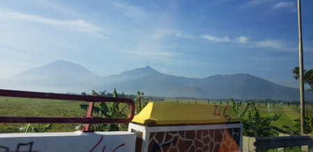 A view of green rice fields and gardens with blue sky and white clouds. You can see the silhouette of a mountain behind him. This view is the view when on the way to Yogyakarta.の写真素材