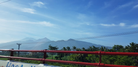 A view of green rice fields and gardens with blue sky and white clouds. You can see the silhouette of a mountain behind him. This view is the view when on the way to Yogyakarta.の写真素材