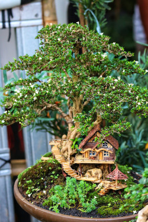 A charming miniature bonsai tree, blossoming with tiny white flowers, supports a quaint wooden treehouse. Mossy details and winding stairs add a touch of magical enchantment.の写真素材