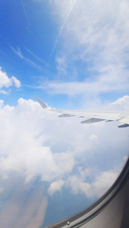 A view from an airplane window showing a portion of the wing against a backdrop of fluffy white clouds and a clear blue sky. The image captures the serenity and vastness of the sky during a flight, offering a tranquil aerial perspective.の写真素材