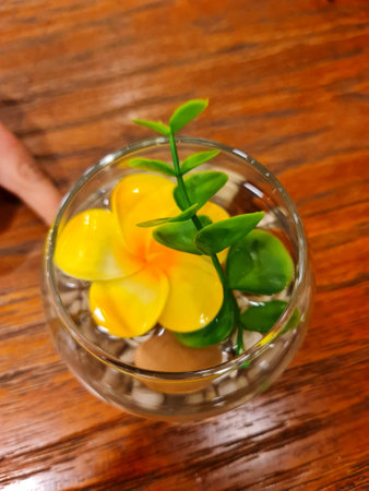 A vibrant yellow plumeria and green sprig float serenely in a glass bowl filled with water and pebbles.  The scene is peaceful and calming.の写真素材