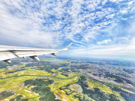 An aerial view from an airplane window shows a vast landscape below a bright blue sky with fluffy clouds. The main focus is the airplane wing, contrasting with the patchwork of green fields and urban areas stretching to the horizon.  The overall mood is tranquil and expansive.の写真素材