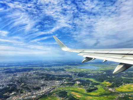 A stunning aerial view from an airplane window showcasing a vast landscape below. Lush green fields and a sprawling city are visible under a vibrant blue sky filled with fluffy white clouds. The airplane's wing is prominently featured in the foreground, adding to the perspective of height and travel.の写真素材