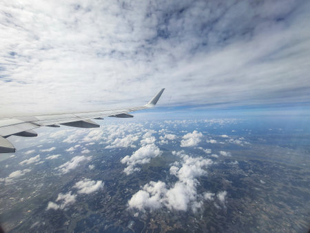 An aerial view from an airplane window shows a vast expanse of fluffy white clouds spread across a patchwork landscape below. The airplane wing is partially visible in the foreground, adding a sense of scale and perspective to the scene. The overall mood is serene and peaceful.の写真素材
