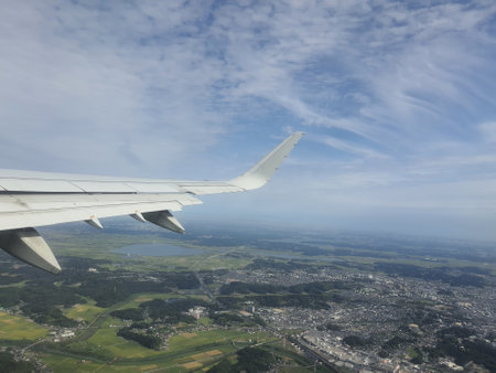 A high altitude aerial view from an airplane window showcasing a wing against a partly cloudy blue sky The landscape below is a patchwork of fields forests and a town. The image evokes a sense of travel and adventure.の写真素材
