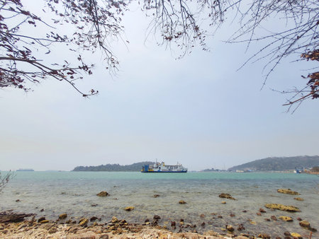 Merak, Indonesia on 21 October 2023. A tranquil seascape featuring a ferry sailing on calm turquoise waters. The foreground shows a rocky shore framed by bare tree branches against a pale sky. The scene evokes a serene and peaceful mood.の写真素材