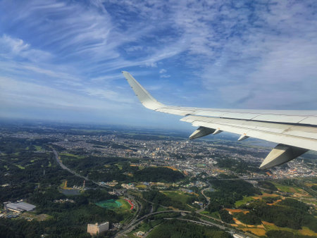 A breathtaking aerial view from an airplane window.  The image showcases a vast cityscape nestled amidst lush green landscapes under a vibrant blue sky. The airplane wing is prominently featured in the foreground, adding to the sense of flight and travel.の写真素材