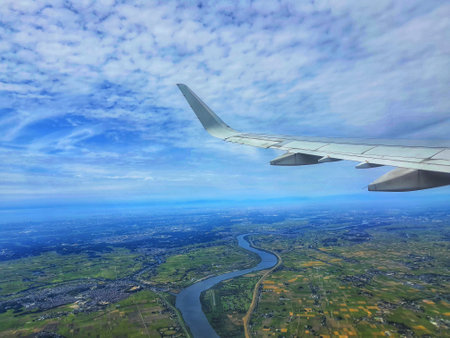 A stunning aerial view from an airplane window showcasing a vast landscape.  A meandering river cuts through patchwork fields, all under a bright blue sky with fluffy clouds. The airplane wing is prominently featured in the foreground, adding a sense of perspective and travel.の写真素材