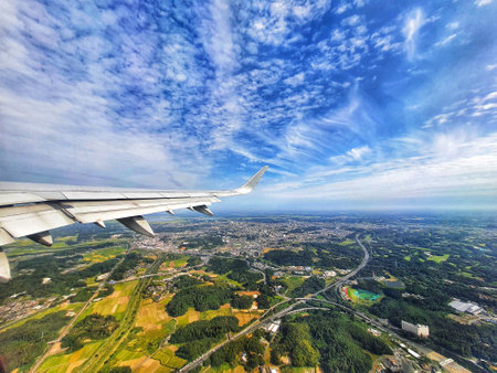 A stunning aerial view from an airplane window showcasing a vast expanse of lush green landscape dotted with roads and buildings under a vibrant blue sky filled with fluffy white clouds. The airplane wing is prominently featured in the foreground, adding to the sense of perspective and height.の写真素材