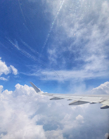 A photograph taken from an airplane window shows a portion of the wing against a backdrop of fluffy white clouds and a vibrant blue sky.  The view is peaceful and expansive, showcasing the vastness of the sky and the feeling of flight. Contrails are visible in the sky, adding an interesting element to the image.の写真素材