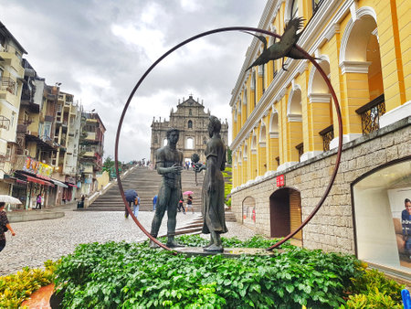Santo Antonio, Macau on 14 Nov 2024. A captivating scene unfolds: Bronze lovers stand framed by a circular arch, overlooking historic Macau ruins and yellow colonial buildings. The rain-washed setting evokes a peaceful atmosphere.の写真素材