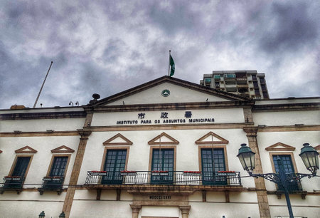 Macau, China on 14 Nov 2024. A grand neoclassical building stands tall under a brooding sky. Dark clouds contrast with the building's pristine white facade, creating a dramatic yet serene atmosphere. Notice the ornate details and iron balconies.の写真素材