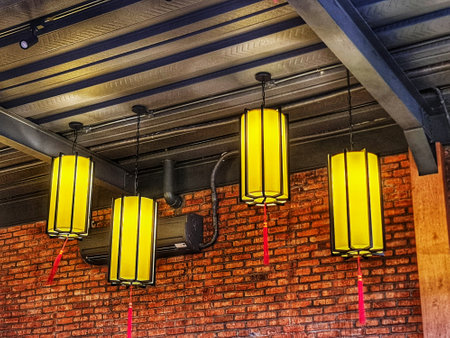 Four bright yellow lanterns hang against a rustic brick wall with visible metal beams and pipes, ideal for interior design and restaurant themes.の写真素材