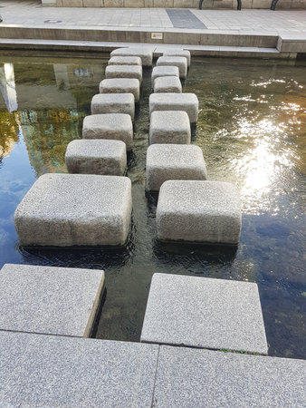 A line of square stepping stones stretches across a clear stream in an urban area offering a tranquil moment for city life and landscape design.の写真素材