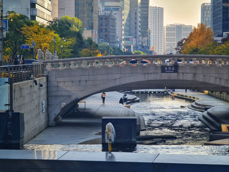 Seoul, South Korea on 11 Nov 2024. A person walks along the paved river walkway, flowing under an arched gray stone bridge in an urban landscape, creating a serene and reflective city scene perfect for travel and tourism marketing.のeditorial素材