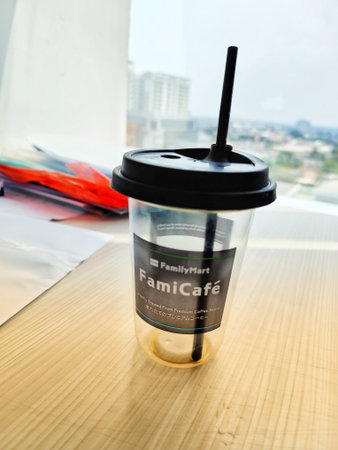 Jakarta, Indonesia on 29 Oct 2025. Clear plastic cup sits with black lid and straw atop a light wooden table near a window in soft daylight, suggesting relaxed drink break for product visualization.のeditorial素材