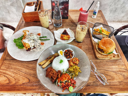 Bogor, Indonesia on 25 Oct 2025. Overhead shot displays a variety of dishes, including a burger, salad, rice plate, fries, and beverages arranged attractively on a rustic wood table surface, perfect for restaurant or food blog promotions.のeditorial素材