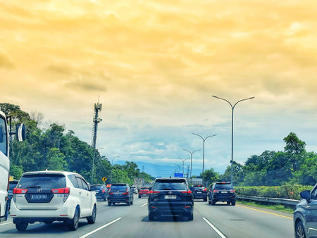 Bogor, Indonesia on 25 Oct 2025. Several modern cars driving on a highway under an overcast yellow sky with trees lining the road and an antenna tower visible, suitable for transportation or travel related marketing.のeditorial素材