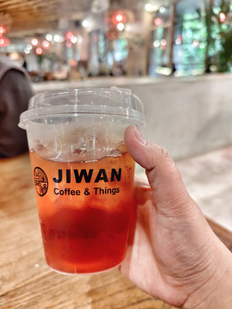 Bogor, Indonesia on 25 Oct 2025. Hand holds a plastic cup filled with reddish-brown iced tea, displayed against a warmly lit indoor cafe background, creating a relaxed and inviting atmosphere perfect for food and beverage marketing.のeditorial素材