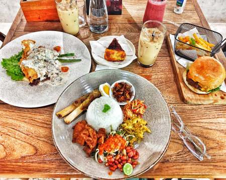 Bogor, Indonesia on 25 Oct 2025. Several plates of assorted food items, including a burger, rice dishes and fried food, are displayed attractively on a textured wooden tabletop with refreshing drinks, creating a casual and inviting atmosphere perfect for のeditorial素材