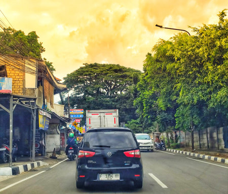 Bogor, Indonesia on 25 Oct 2025. Dark car moving on a paved road lined with buildings and lush trees under an orange tinted sunset sky, creating a peaceful urban travel scene, ideal for transportation or travel related content.のeditorial素材