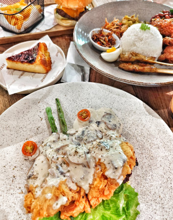 Bogor, Indonesia on 25 Oct 2025. Various dishes like fried chicken with creamy mushrooms, rice, burger, french fries and cake are presented in a close-up shot on a wooden table, ideal for food blog or restaurant related advertising.のeditorial素材