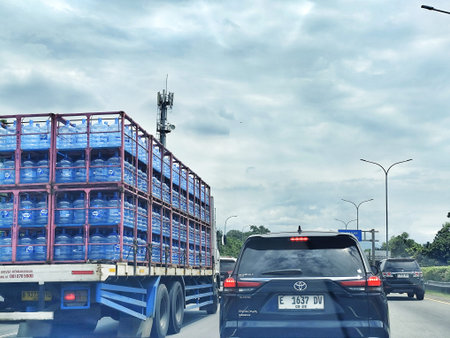 Bogor, Indonesia on 25 Oct 2025. Truck hauling stacked blue water bottles is driving on the highway with other cars under a cloudy sky, creating a sense of transportation and industry suitable for supply chain or travel marketing.のeditorial素材