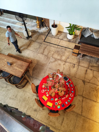 Bogor, Indonesia on 25 Oct 2025. An indoor scene from a high angle shows a man standing near a wooden table, with a red tablecloth-covered round table surrounded by chairs and potted plants, creating a casual and inviting atmosphere suitable for interior のeditorial素材